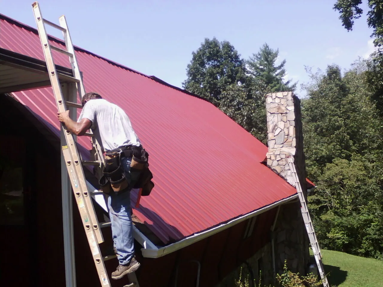 Roofer on a ladder beside a red metal roof and stone chimney — Soffit & Fascia Repair by Roofing Brothers in Thorsby
